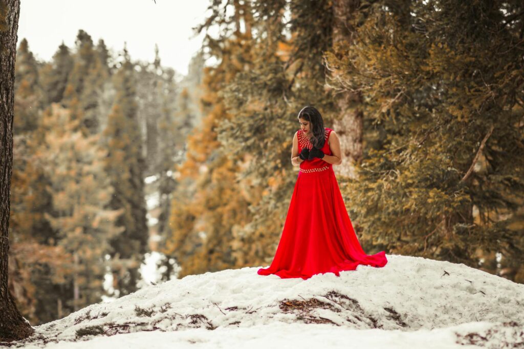Woman in Red Dress Standing on Snow Covered Ground Surrounded by Trees